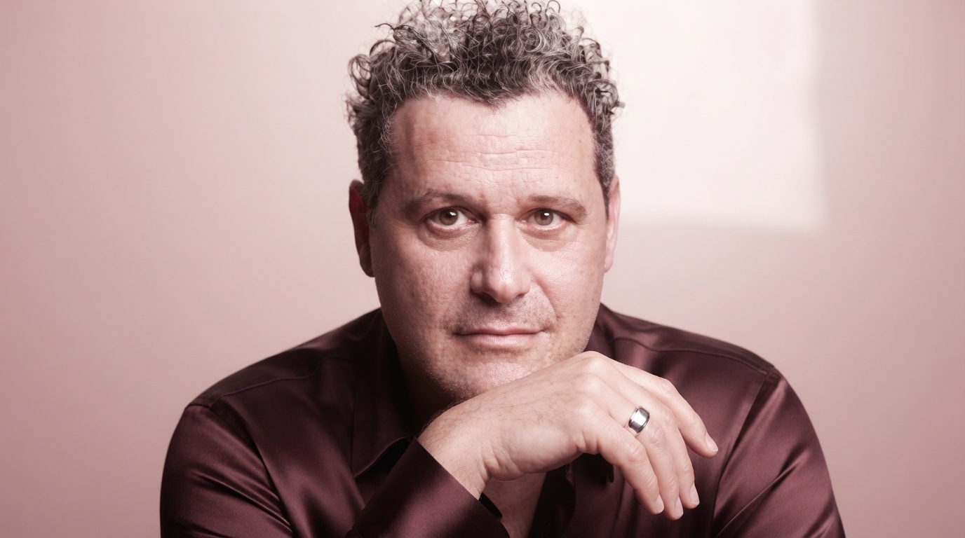 Isaac Mizrahi in a studio setting, surrounded by colorful fabrics and design sketches, looking thoughtfully at a piece of clothing.